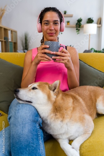 A Hispanic woman relaxes on a sofa with her dog and drinks from a mug