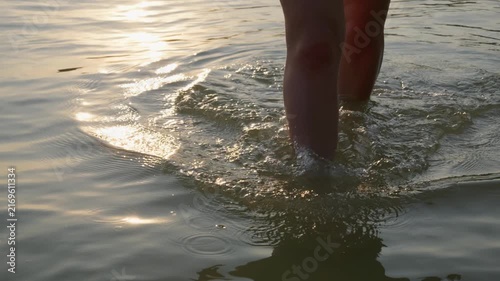 Bare legs wade through shallow, calm lake water during a peaceful sunset in slow motion. Circular ripples spread gently across the surface, reflecting bright golden evening light.
