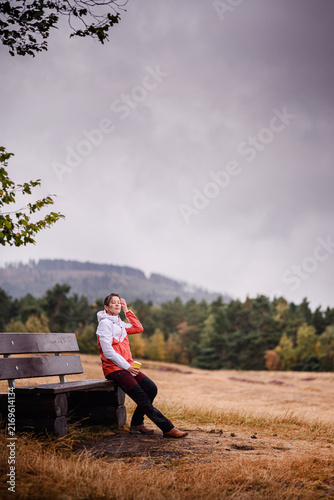 Fotografie Hiker in red jacket leaning against wooden bench holding coffee cup and enjoying fresh air on gloomy autumn day