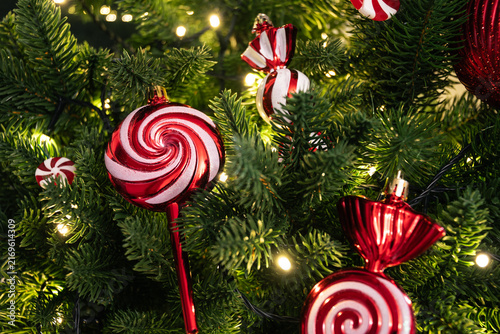 Red and white candy shaped ornaments hanging on green Christmas tree with warm glowing lights. Festive holiday decoration and Christmas background.