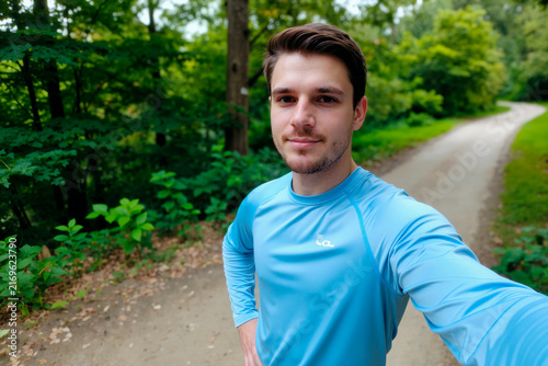 Portrait of Caucasian young adult man posing on forest trail, holding smartphone for fitness selfie and looking at camera. Ideal for wellness, outdoor exercise, active lifestyle marketing