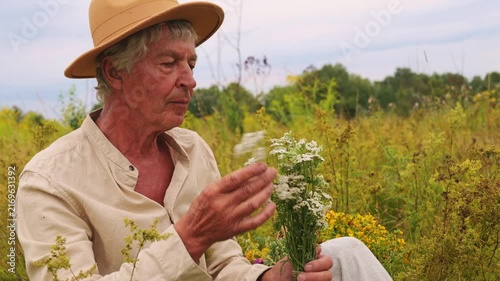 A man sits in a grassy field holding a bunch of white flowers. He inspects the herbs carefully. The day is cloudy and the area is filled with various plants