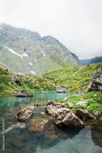 Canvastavla Hikers rest beside clear Blue Abudelauri Lake beneath cloud-covered rocky peaks