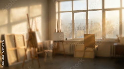 Empty artist studio features wooden easels and blank canvases positioned near a large window. Soft golden sunlight streams through the glass, creating a dreamy atmosphere with a blurred focus.