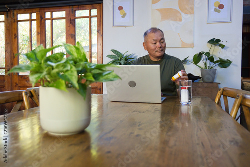 Senior Asian man sitting at dining table holding pill bottle beside laptop and glass, copy space