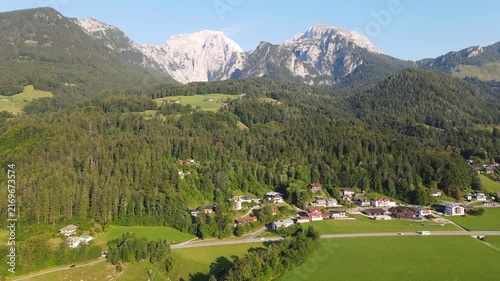Aerial drone view of the peaceful alpine countryside near Schönau am Königssee in Bavaria, Germany, with lush green meadows, traditional Bavarian houses and majestic mountain ranges in the background.