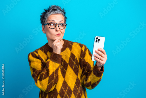 Skeptical senior woman with smartphone wearing glasses and argyle sweater against blue background