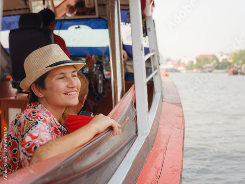 Female tourist in elegant summer dress and straw hat enjoys scenic boat ride along Chao Phraya River in Bangkok. Woman looks at waterfront view and enjoys refreshing breeze.