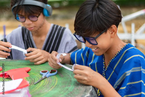 Two happy teenage boys in traditional shirts smiling and holding paper rolls for an art craft project together at an outdoor green table.