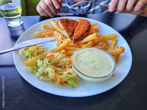 Child hands holding cutlery to eat a plate of chicken schnitzel, french fries, fresh salad, and sauce at a restaurant table.