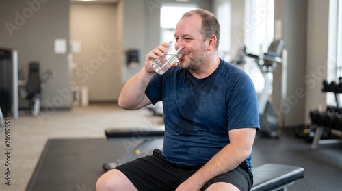 Sweaty man drinking a glass of cold water to rehydrate after an intense workout at the gym with sports club cool down hydration recovery