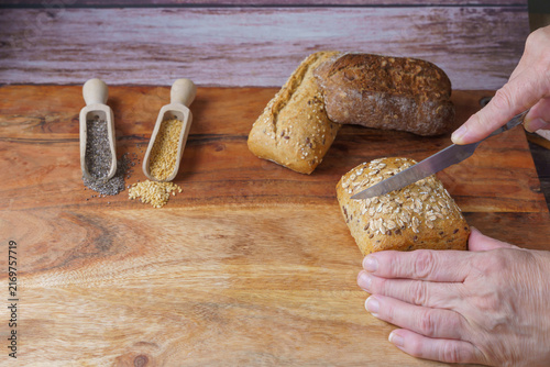 Hands cutting a fresh whole grain bread roll with oats, flax, and chia seeds on a rustic wooden board, preparing healthy meal