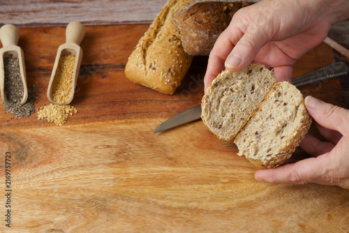 Hands holding a freshly sliced piece of seeded whole grain bread over a wooden cutting board with chia and flax seeds