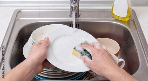 First person point of view of person washing a white ceramic dinner plate with a soapy yellow sponge under running tap water in a kitchen stainless steel sink