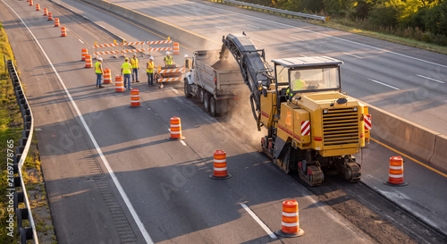 Aerial view of a road milling machine stripping damaged asphalt pavement from a highway roadway with construction crew and safety cones in place