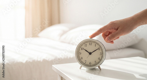 Hand reaching out to silence classic white alarm clock on bedside table next to bed in bright morning bedroom