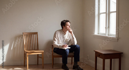 Pensive thoughtful man seated on a simple wooden chair inside a minimalist sparsely furnished room looking anxiously out the window with hand on chin