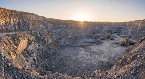 Wide panoramic landscape view of a massive open pit rock quarry with industrial machinery under bright golden sunset skies.