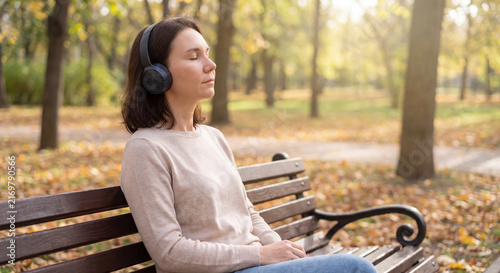 Young woman sitting on a wooden bench in an autumn park with closed eyes listening to peaceful music through modern headphones enjoying fall nature
