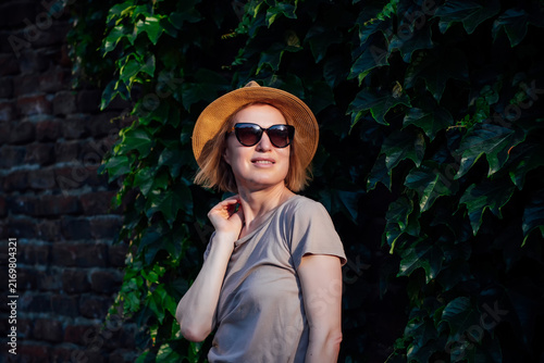 Stylish young woman in sunglasses and straw hat posing by lush ivy wall in warm evening light. Summer fashion portrait with natural greenery, relaxed mood and copy space