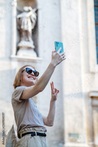 Smiling female traveler taking a selfie with a smartphone in front of historic architecture. Sunny vacation moment blending tourism, technology and joyful memories