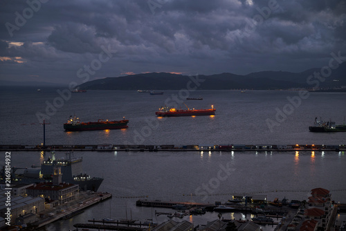 Tankers anchored in the Bay of Gibraltar at dusk beneath dramatic storm clouds, with harbour lights and reflections across the calm waters of the Strait of Gibraltar