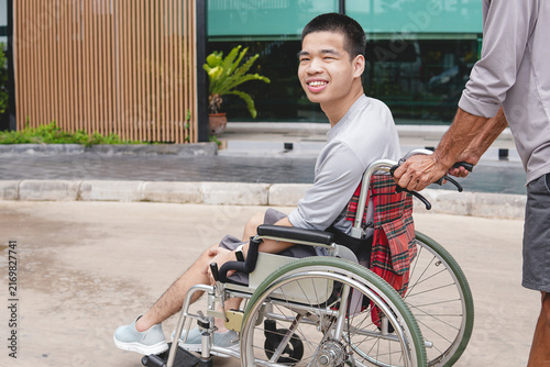 Fotografie Happy young Asian man in a wheelchair looking at camera with a smile while pushed by caregiver outdoors, concept of positive attitude, accessibility, and inclusion