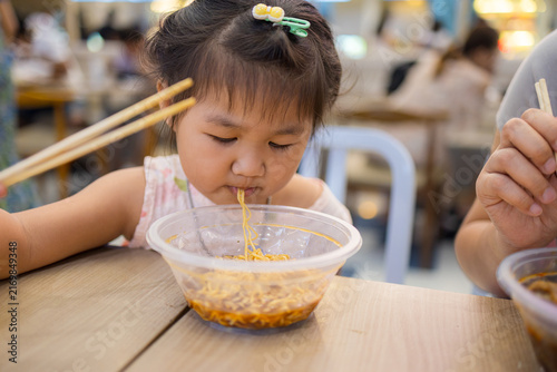 Konstfotografi Cute Asian Girl Eating Instant Noodles with Chopsticks at a Restaurant,Close-up of an adorable little Asian girl enjoying a bowl of delicious instant noodles with chopsticks in a casual restaurant