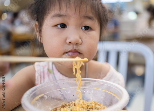 Photography Cute Asian Girl Eating Instant Noodles with Chopsticks at a Restaurant,Close-up of an adorable little Asian girl enjoying a bowl of delicious instant noodles with chopsticks in a casual restaurant