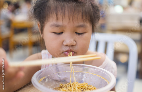 Картината върху платно Cute Asian Girl Eating Instant Noodles with Chopsticks at a Restaurant,Close-up of an adorable little Asian girl enjoying a bowl of delicious instant noodles with chopsticks in a casual restaurant