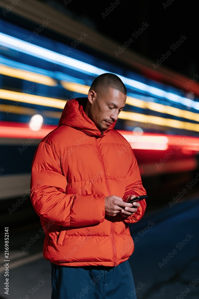 Fototapeta premium Man in red puffer jacket using smartphone at night by moving train with light trails for urban commute portrait and neon background