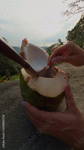 Hands Scoop Tender Coconut Flesh from an Opened Green Coconut on a Rustic Table, Framed by Forested Hills and Distant Water, Showing Fresh Tropical Food Preparation Vertical video.