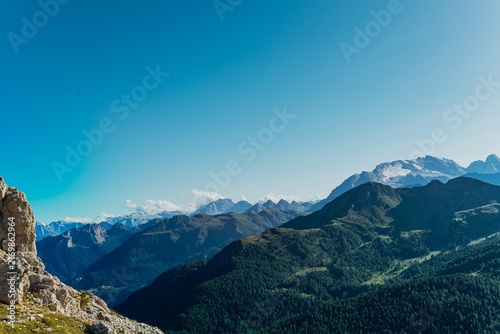 Layered green ridges fade into hazy blue Dolomite peaks under a bright morning sky. Serene mountain layers and misty depth for wellness, meditation and travel background use