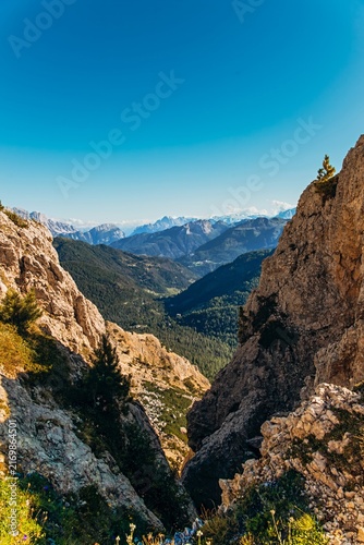 Steep rocky gorge frames a distant valley and blue mountains seen from a high hiking path. Vertigo and scale of alpine hiking for adventure travel, trekking gear and outdoor marketing