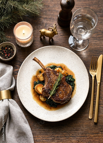 Roasted lamb chop with rosemary, vegetables, candlelight, wine glass, cutlery, and evergreen branches arranged on a rustic wooden dining table.