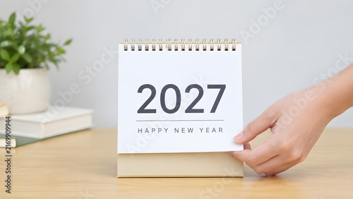 A hand turning the page of a desk calendar showing the year 2027 for a happy new year celebration in a bright home office setting on a wooden table with a small green plant