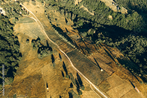 Aerial view of sunlit mountain meadows, winding dirt roads and dense forest creating a peaceful rural landscape.
