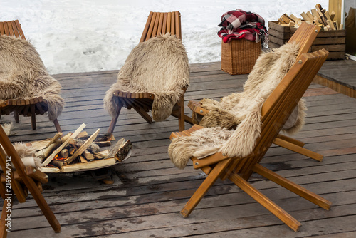 An outdoor seating area in winter. Wooden chairs covered with fur blankets surround a fire.