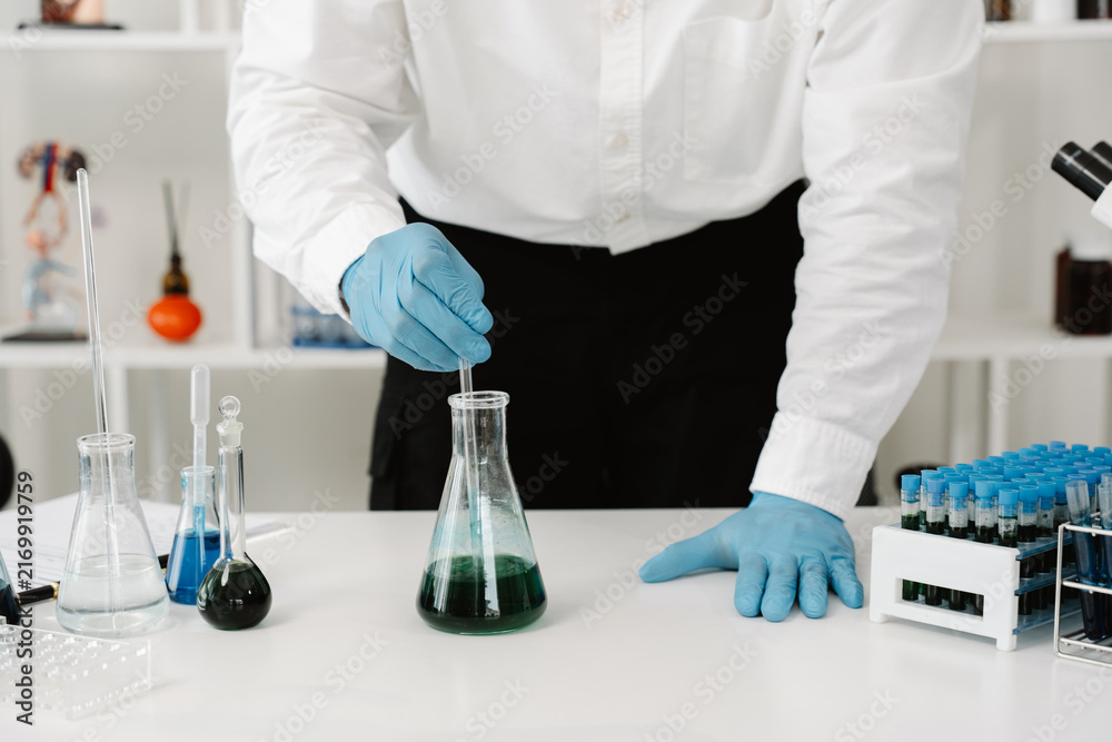 Fototapeta premium Scientist Mixing Liquid in Flask: A scientist in a lab coat and blue gloves carefully mixes green liquid in a flask, surrounded by laboratory equipment and test tubes.