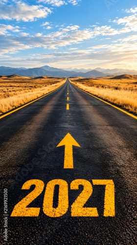 Year 2027 painted on an empty asphalt road leading into the distance with a bright yellow arrow pointing forward towards the horizon under a beautiful sunset sky and open landscape
