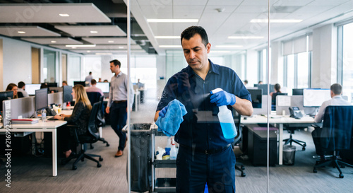 Male janitor cleaning a glass partition with spray and cloth in a busy modern corporate office