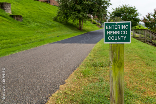 View of the Entering Henrico County sign on a wooden post beside a paved path and grassy hill in Henrico, Virginia, United States.