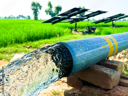Clear water flowing from a solar-powered tube well into a lush green paddy field, with solar panels visible in the background