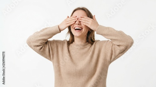 Happy young woman covering eyes with hands and smiling broadly while wearing a casual beige shirt in a joyful mood