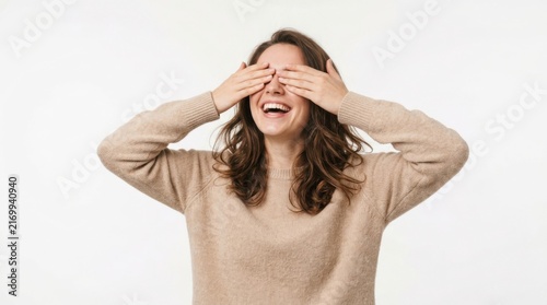 Happy young woman wearing a beige sweater covering her eyes with her hands and smiling broadly in a playful mood