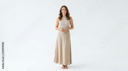 A beautiful young woman with brown hair standing and smiling wearing a simple long beige summer dress in a bright room