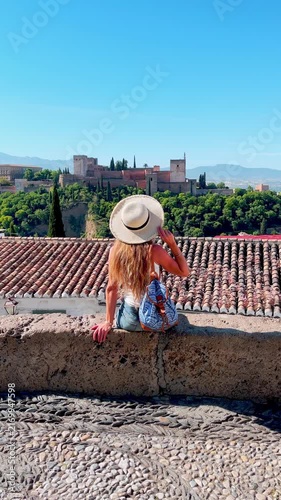 woman in straw hat sitting on rooftop overlooking alhambra palace and red tiled roofs in granada spain, vertical footage