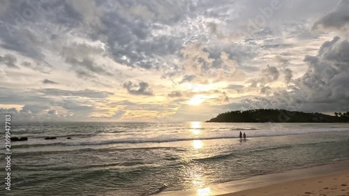 Wide shot of a beautiful sunset on a Sri Lanka beach showing people entering the sea, soft yellow sand, dramatic clouds, and distant cliffs covered with vegetation at nighttime.