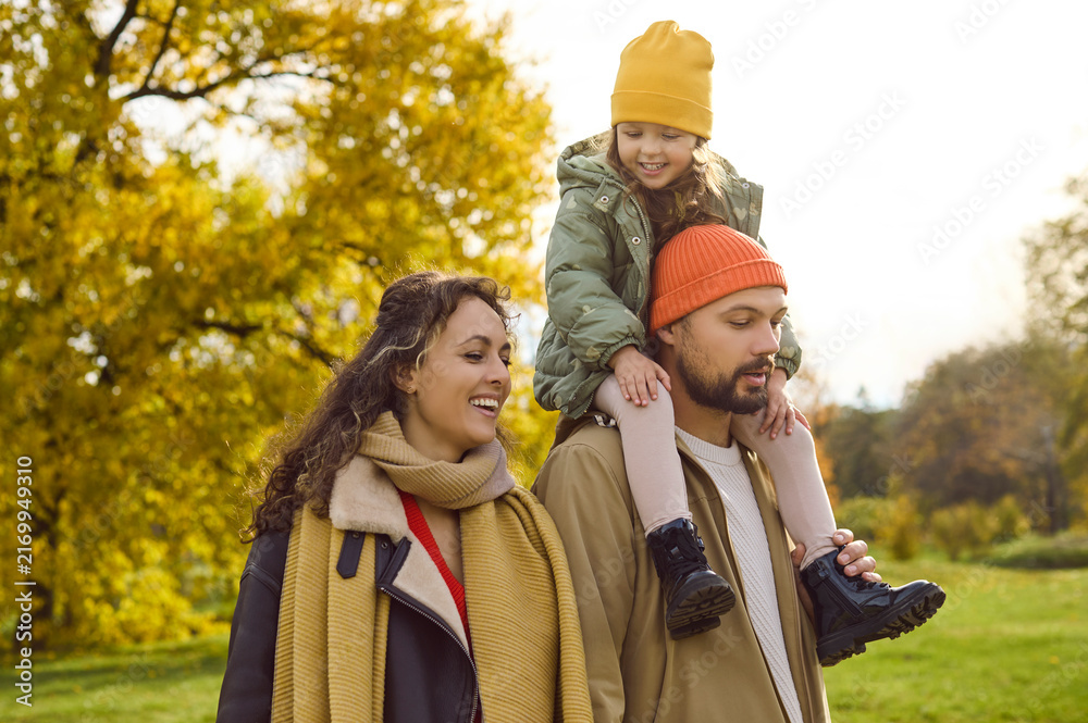 Fototapeta premium Happy family enjoys autumn walk in park. Parents in warm layers smile as their daughter rides on dads shoulders among golden leaves, sharing laughter. A warm scene of love and happiness.
