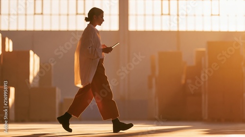 Small business owner walking through a warehouse, holding a tablet, with large cardboard boxes stacked in the background and sunlight streaming through windows